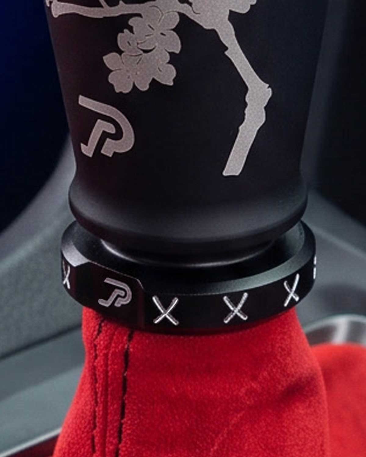 A close-up of a black anodized shift knob and matching boot retainer with white cherry blossom engravings, installed on a red leather gear shift boot with black stitching.
