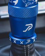 A close-up of a blue anodized shift knob and matching boot retainer with white koi fish laser engravings, installed on a black leather gear shift boot with white stitching.
