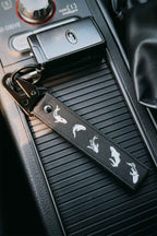 A black embroidered car keychain strap with white koi fish silhouettes, attached to a Subaru STI key ring and placed on a car center console.