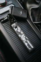A black embroidered car keychain strap with white sakura cherry blossom silhouettes, attached to a Subaru STI key ring and placed on a car center console.