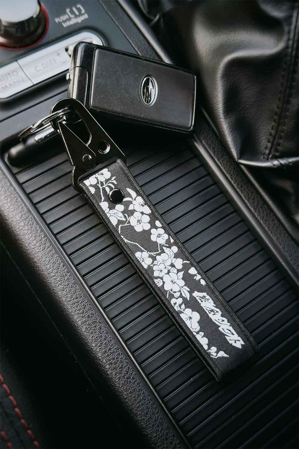 A black embroidered car keychain strap with white sakura cherry blossom silhouettes, attached to a Subaru STI key ring and placed on a car center console.