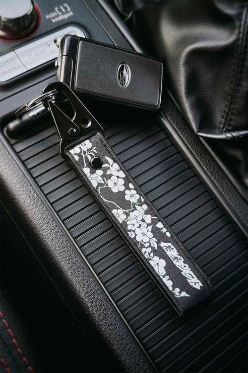 A black embroidered car keychain strap with white sakura cherry blossom silhouettes, attached to a Subaru STI key ring and placed on a car center console.
