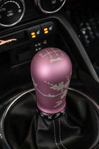 A pink manual gear shift knob with sakura engravings, Miata logo, and a 6 speed shift H pattern, paired with a matte black boot retainer with slot design, installed in a Mazda Miata's center console.