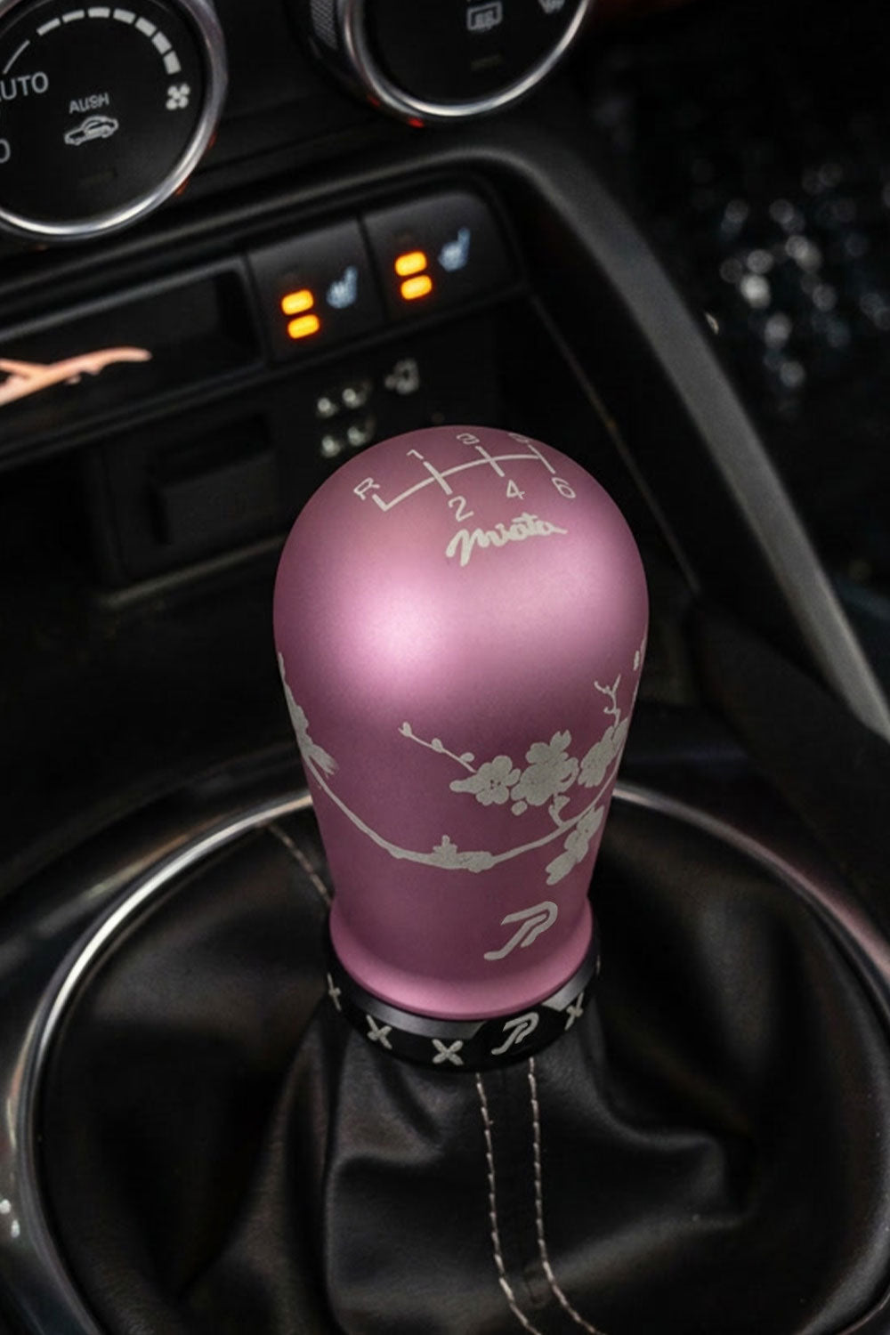 A pink manual gear shift knob with sakura engravings, Miata logo, and a 6 speed shift H pattern, paired with a matte black boot retainer with slot design, installed in a Mazda Miata's center console.