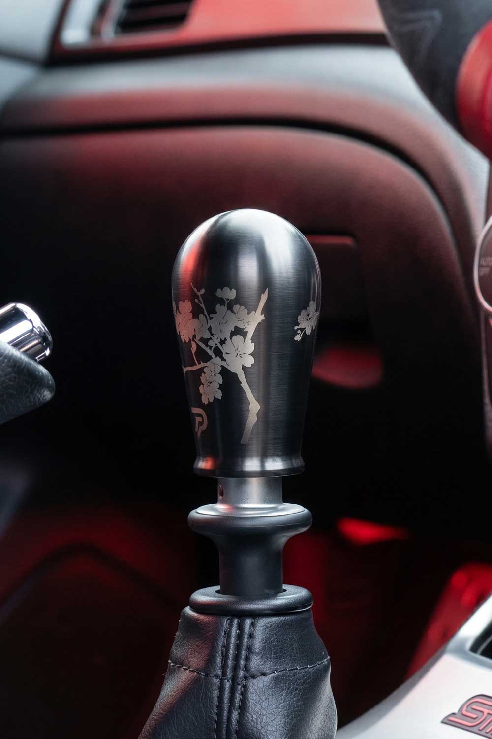 A close-up of a custom black metal manual gear shift knob with an etched cherry blossom branch design, set inside a car's console with red ambient lighting.
