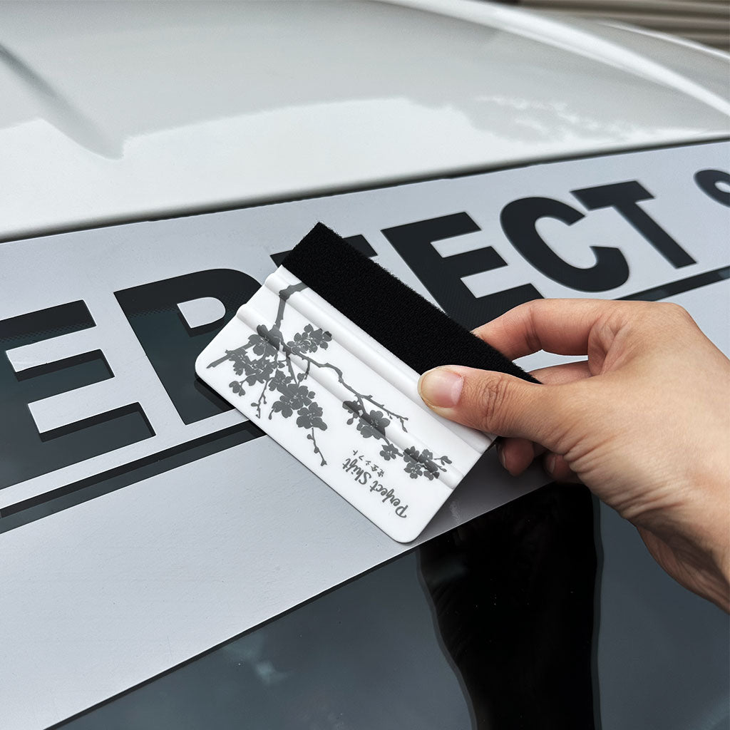 A hand holding a banner sticker application squeegees with cherry blossom pattern and Perfect Shift logo and demonstrate on a grey banner 