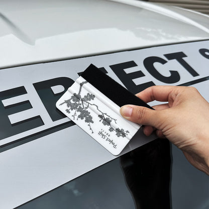 A hand holding a banner sticker application squeegees with cherry blossom pattern and Perfect Shift logo and demonstrate on a grey banner 