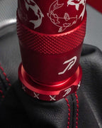 A close-up of a red anodized shift knob and matching red boot retainer with white koi fish laser engravings, installed on a black leather gear shift boot with red stitching.
