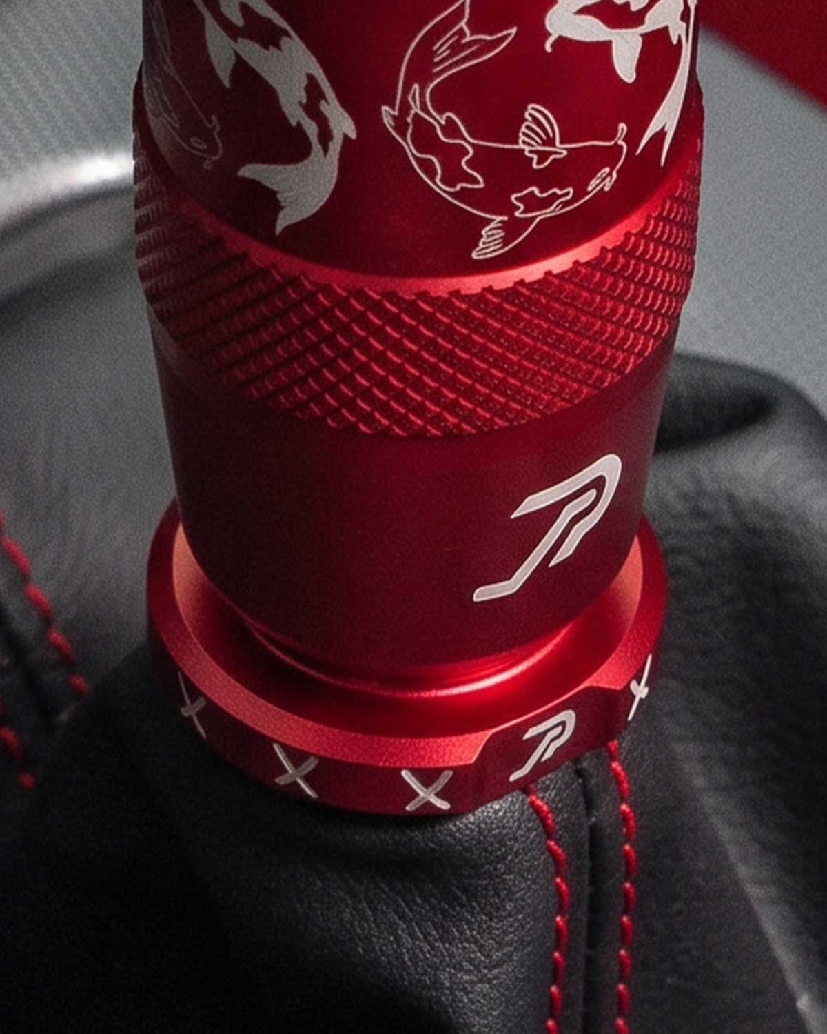 A close-up of a red anodized shift knob and matching red boot retainer with white koi fish laser engravings, installed on a black leather gear shift boot with red stitching.