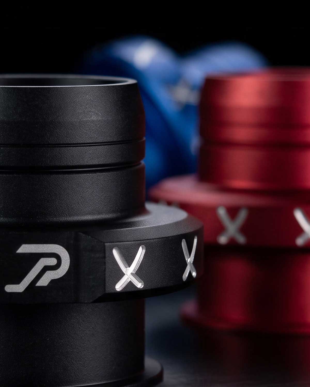 Close-up of a black shift boot retainer with silver logo and slot markings, with a blurred red and blue version in the background.