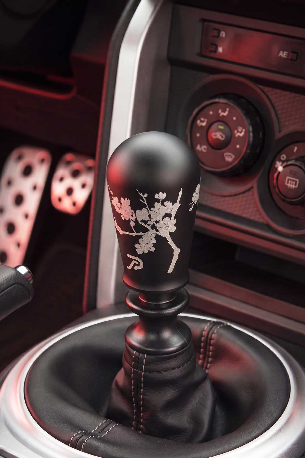 A close-up of a matte black manual gear shifter with a cherry blossom branch engraving, installed in a GT86's center console with red ambient lighting.