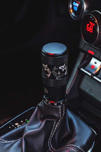 A close-up of a black shift knob with an engraved koi fish design, mounted on a black leather gear shift boot inside an automatic Toyota GR86's console.