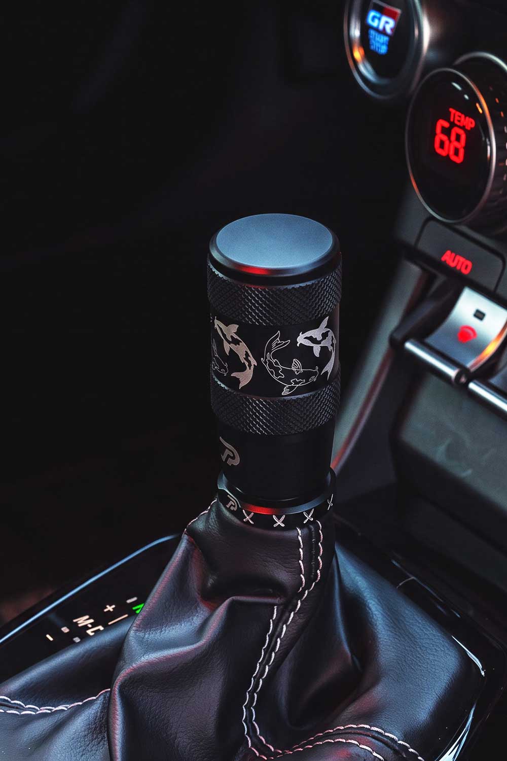 A close-up of a black shift knob with an engraved koi fish design, mounted on a black leather gear shift boot inside an automatic Toyota GR86's console.