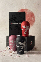 Three tear-drop shaped JDM car shift knobs in black, red, and pink, each decorated with a white crisp cherry blossom engraving and a Perfect Shift logo, arranged in front of black branded boxes on a textured beige surface with scattered petals and a red Japan rising sun backdrop.