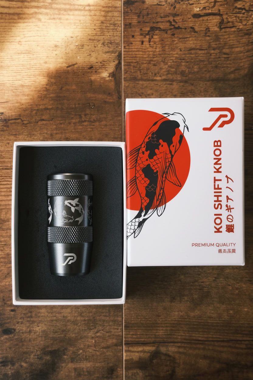 A dark gun metal Koi Shift Knob presented in an open box next to its product packaging box with red and black koi fish graphics and Japanese characters, reading 'premium quality shift knob', and set on a rustic wooden background.