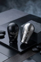 Two stainless steel shift knobs displayed on a dark stone slab: one brushed black with cherry blossom engraving, and one silver with matching engraving, set with dark stones against a moody dark background.