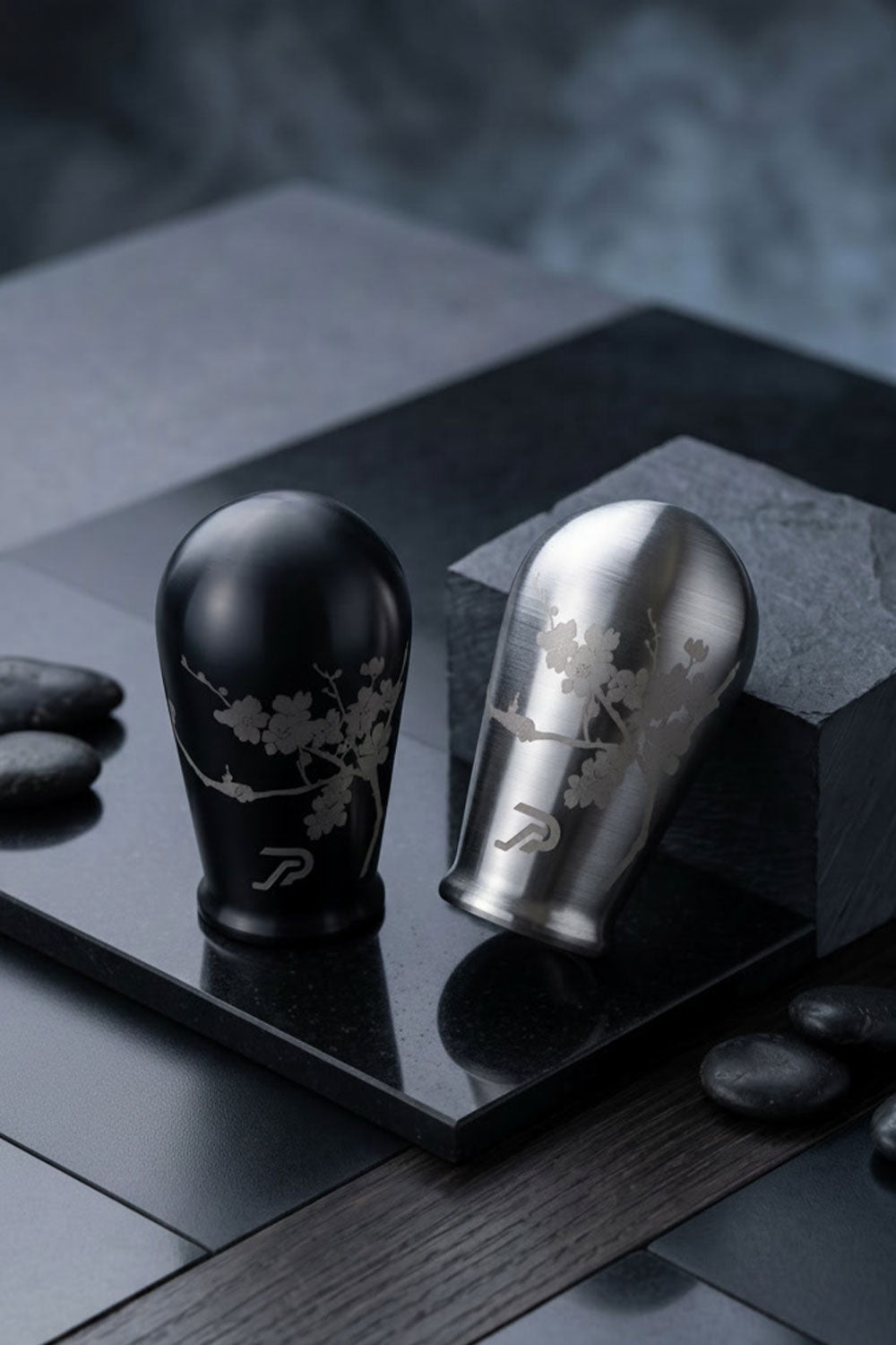 Two stainless steel shift knobs displayed on a dark stone slab: one brushed black with cherry blossom engraving, and one silver with matching engraving, set with dark stones against a moody dark background.