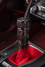A close-up of a black stainless steel shift knob with white koi fish engraved graphics, installed in a Honda Type-R with red suede gear shift boot.