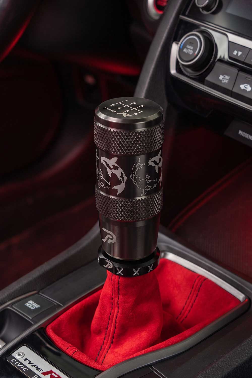 A close-up of a black stainless steel shift knob with white koi fish engraved graphics, installed in a Honda Type-R with red suede gear shift boot.